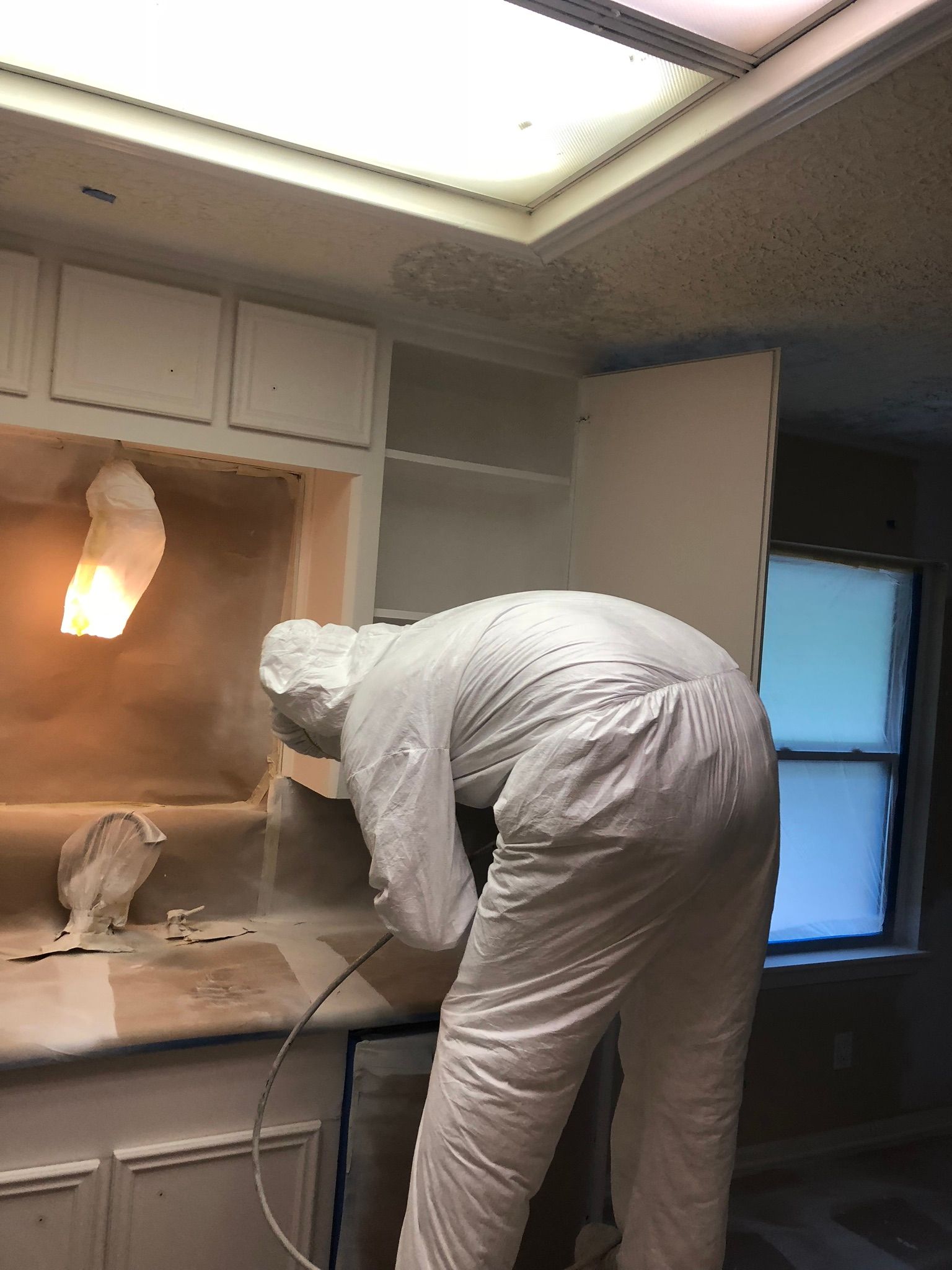 A man in a white suit is spray painting a cabinet in a kitchen.